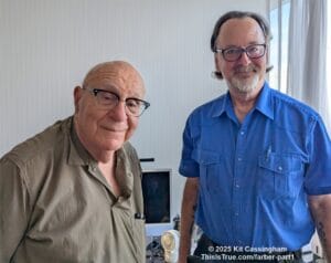 Two men with glasses stand indoors, smiling at the camera. On the left is Dave Farber, in a light brown shirt, and on the right, Randy Cassingham in a blue shirt. A computer and a small fan are visible in the background.