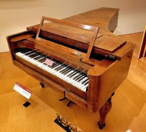 A vintage wooden grand piano with ornate legs is displayed in a museum. The piano lid is partially open, and informational signs are placed nearby on the wooden floor.