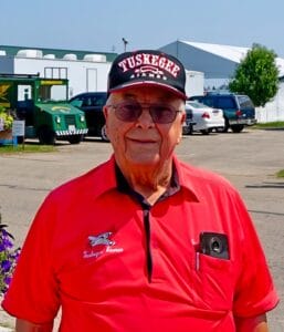 An elderly man wearing a red Tuskegee Airmen cap and matching polo shirt stands outdoors, smiling. He has glasses and a pen in his shirt pocket, with vehicles and buildings in the background.