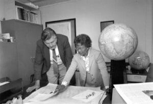 A woman and a man stand in an office, examining computer-generated satellite data spread across a desk. There are two large globes available for reference.