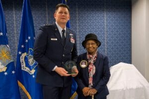 A uniformed Air Force general holds an award while standing next to an elderly woman in a hat and glasses. Air Force flags are visible in the background. Both are posing and smiling for the photo.