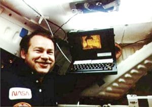 An astronaut in a NASA uniform smiles while holding a laptop inside a space shuttle. The laptop screen displays an image, and the background shows the interior walls and equipment of the spacecraft.