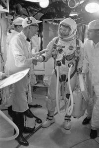 An astronaut in a spacesuit shakes hands with a man in a lab coat and cap, surrounded by others in similar attire inside a spacecraft or control room, preparing for a mission.