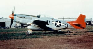 A World War II-era silver P-51 Mustang fighter plane with a red tail and American star insignia is parked on a grassy field near hangars.