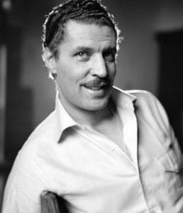A man with short, dark hair and a mustache smiles while leaning back in a chair. He is wearing a light-colored, unbuttoned collared shirt. The photo is in black and white, and the background is softly blurred.