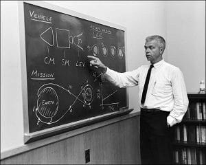A man in a white shirt and tie points to a chalkboard with diagrams of a spacecraft, mission path from Earth, and labeled escape velocities, in a conference room.