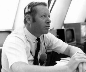 A man in a short-sleeve shirt, tie, and headset sits at a desk with papers, appearing focused. In the background, there is a vintage television and large windows letting in light.