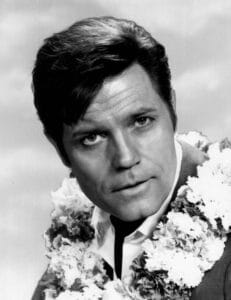 Black and white portrait of Jack Lord with dark hair, wearing a suit and tie, adorned with floral leis around his neck, looking directly at the camera with a serious expression.