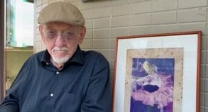 Jaguar as an older man wearing glasses, a beige flat cap, and a dark shirt sits next to a framed artwork of a ballerina in a pink tutu against a tiled wall.