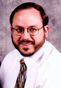 A man with short brown hair, a beard, and glasses is smiling at the camera. He is wearing a white shirt, patterned tie, and is posed in front of a gray textured background.