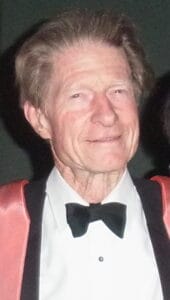 An older man with light skin and gray hair wearing a black bow tie, white shirt, and a jacket with pink satin lapels, smiling at the camera.