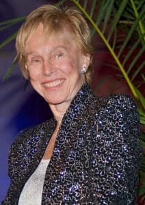 A smiling woman with short blond hair wears a sparkly black and silver jacket, pearl earrings, and a necklace, standing in front of green foliage.