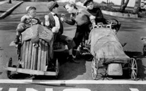 Four children in early 20th-century clothing sit in handmade wooden soapbox derby cars at a starting line. One child (Kibrick) stands with a raised fist, confronting another, while the others watch.