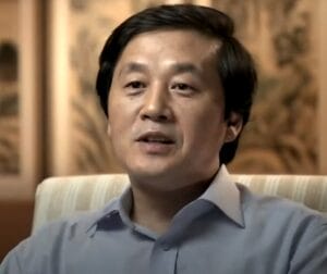 A man with short dark hair wearing a light blue collared shirt sits in a striped chair, speaking, with a softly blurred, warm-toned background featuring artwork.
