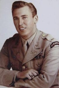A young man in a light-colored U.S. Army dress uniform with military badges, ribbons, a Ranger patch, and crossed rifles insignia, smiles while sitting with his arms crossed on a table.