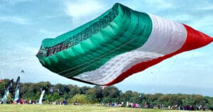 A gigantic kite with green, white, and red stripes flies close to the ground at an outdoor event, with people and tents visible in the background on a grassy field.