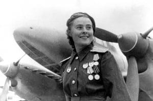 A smiling woman in a military uniform with medals stands in front of a large propeller aircraft, with clouds in the sky behind her.