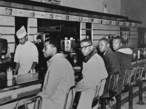 Four young Black men sit at a lunch counter while a Black server works behind it. The men appear calm and determined, participating in a peaceful sit-in protest at a segregated diner in the 1960s.