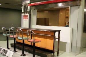 A lunch counter with four metal stools, part of a museum exhibit, is displayed behind a glass barrier. There is a historical information sign and a banner about the Greensboro Lunch Counter sit-in nearby.