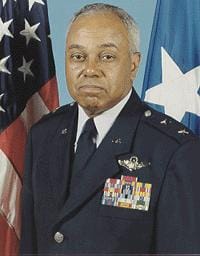 A man in a decorated military uniform stands in front of American flags and a blue flag with a white star. He has short gray hair and is looking at the camera with a neutral expression.