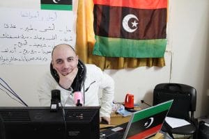 A man in a white jacket sits at a desk with computer monitors and Libyan flags. A large Libyan flag and a poster with Arabic text hang on the wall behind him. He is smiling and resting his chin on his hand.