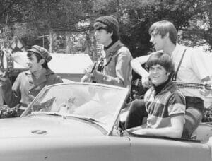Four young men sit and play musical instruments in and on a convertible car. One holds a tambourine, another a guitar, one a bass, and the fourth is sitting on the car’s back, smiling. Trees and houses are visible in the background.