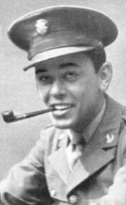 A man in a military uniform and cap smiles at the camera while holding a pipe in his mouth. The image is black and white and from World War II.