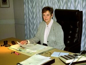 Rimington in a light gray suit sits at a desk covered with papers, a calculator, glasses, and office supplies. She faces the camera, holding a document, with a neutral expression in a professional office setting.