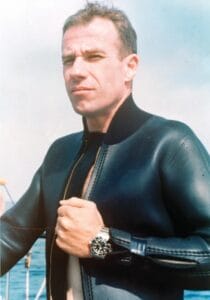 A man wearing a black wetsuit and a wristwatch stands outdoors, with his hand holding the front zipper. The background shows a partly cloudy sky and some water, suggesting a marine setting.