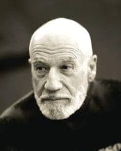 An older man with a bald head and white beard looks thoughtfully to the side. He is wearing a dark shirt and the background is blurred. The photo is black and white.