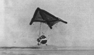A small delta-wing hang glider over a flat landscape, with distant hills visible in the background. The glider is carrying a space capsule.
