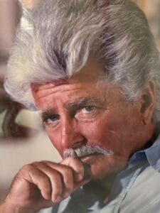 A man with thick, wavy gray hair and a mustache gazes intently at the camera, resting his hand near his mouth. He wears a light blue shirt and has a thoughtful, serious expression.