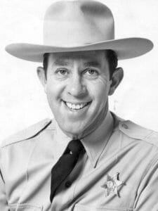 A man dressed as a generic sheriff smiles at the camera, wearing a wide-brimmed hat, a uniform shirt with a star-shaped badge, and a dark tie. The image is in black and white.