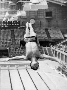 A male diver in midair, upside down with arms outstretched, above an indoor swimming pool with empty spectator seats in the background.