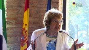 An older woman speaks passionately at a lectern with microphones. Behind her are the Spanish flag and another flag. She is indoors, near a brick wall and a window, with a logo in the upper right corner.