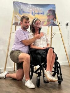 Hugo kneels beside a Leah in a wheelchair, holding a microphone for her. Behind them is a banner that reads Her ALS Story Warrior Weekend with an illustrated background.