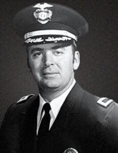 A man in a police dress uniform, with a peaked cap featuring a badge, dark jacket with insignia on the shoulders, and a tie, poses for a formal black and white portrait against a plain background.