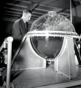 A man in a suit stands behind a large, transparent, spherical object mounted on a metal base, inside an industrial or laboratory setting.