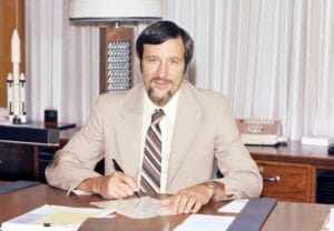 A man with brown hair and a beard, wearing a beige suit and striped tie, sits at a desk with papers and a pen in hand. Behind him is a model rocket.
