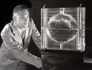 A man in a suit looks at a small, spherical scientific apparatus enclosed in a gridded, transparent case. The setting is indoors, with a dark background.