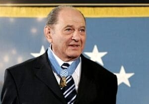A man in a suit and striped tie wears a Medal of Honor around his neck, standing in front of a blue background with white stars.