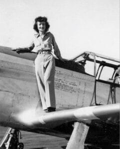 A woman in a button-up shirt and trousers stands smiling on the wing of a military aircraft from the World War II era. The photo is in black and white.
