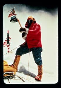 A climber in a red jacket and yellow boots stands on the summit of Mount Everest, holding an ice axe with attached flags. Ropes are visible, and wind or mist swirls in the background. The climbers face is covered by goggles and a mask.